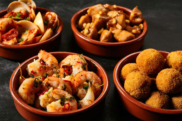 Four terracotta bowls display various dishes. Shrimp garnished with herbs, saucy clams, savory chicken pieces, and golden-brown croquettes rest against a dark, textured surface.